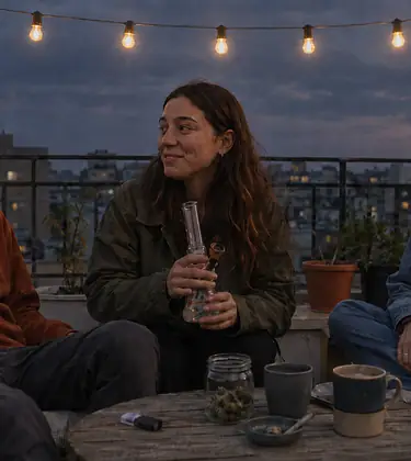 Three friends laughing around a low table on a city balcony at dusk, one holding a clear glass water pipe beside a jar of flower