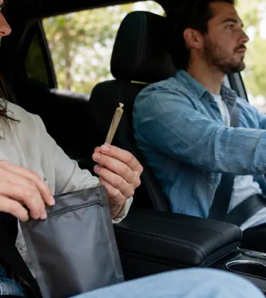 Passenger holding an unlit cannabis pre-roll and resealable pouch inside a car while the driver focuses on the road.
