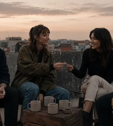 Four young friends on a city rooftop at dusk, one passing a joint sideways mid-conversation