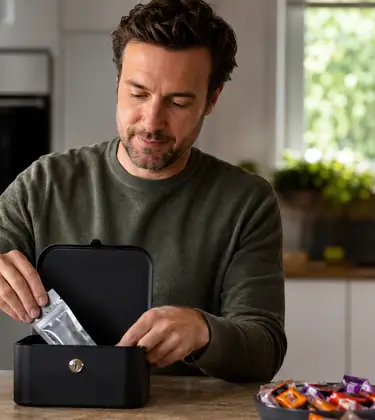 Man placing cannabis edibles into a lockbox beside Halloween candy and a pumpkin in a home kitchen.