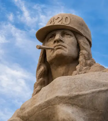 Stone monument wearing a carved stone 420 cap with a carved stone blunt in its mouth against a blue sky.