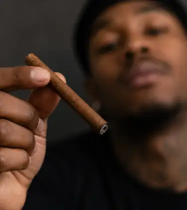 Professional athlete in dark sportswear holding a blunt in the foreground, with his face softly blurred in the background.