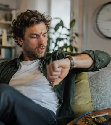 Man relaxing on a couch with a joint, checking his watch while a blurred wall clock suggests cannabis time distortion.