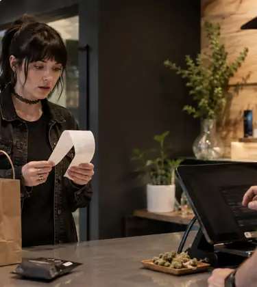 A surprised customer reads a long receipt at a modern cannabis dispensary checkout while a budtender operates the register.