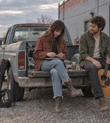 Two young friends sitting on the tailgate of a pickup truck behind a small music venue, one rolling a joint on a tin, the other holding an acoustic guitar