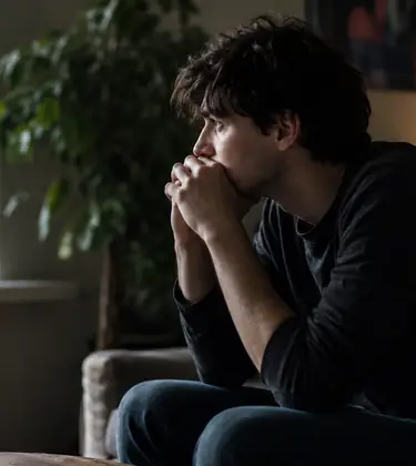 Young man sitting on a sofa by a window, looking anxious and deep in thought in a dimly lit living room.