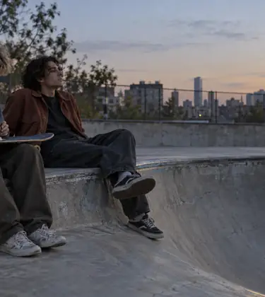Two young skaters sitting on the coping of an empty concrete bowl at dusk, one grinding flower on an upturned deck across his knees, boards and a backpack beside them