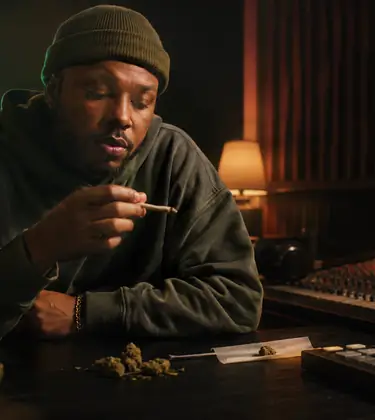 A hip-hop artist holding a joint in a dimly lit recording studio, surrounded by a mixing console, beat machine, and cannabis buds.