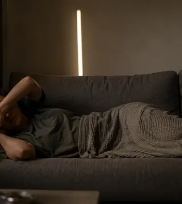 Woman lying on a sofa in a dim room with the curtains drawn and one arm across her eyes.