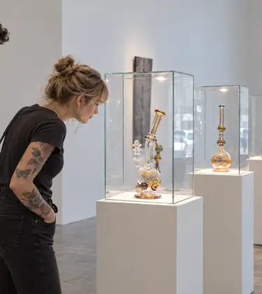 Two young gallery visitors looking at an elaborate colored glass water pipe on a lit plinth, more glass pieces on plinths behind them