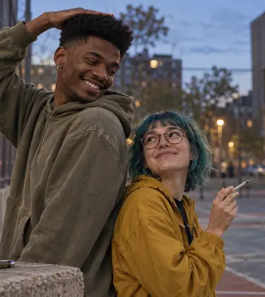 Two young friends of very different heights compare heights beside a city basketball court at dusk, one holding a joint