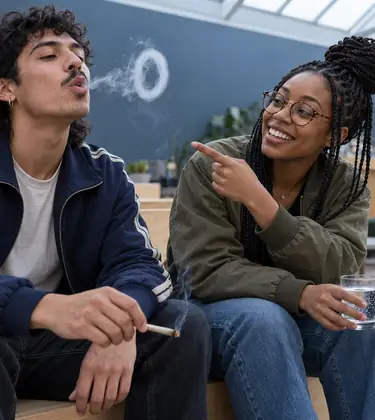 Two friends on a lounge bench, one blowing a smoke ring from a joint while the other points at it and grins