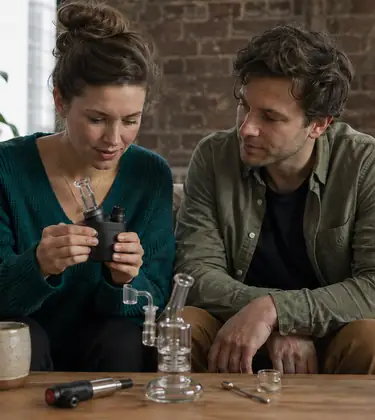 Two young friends on a loft sofa examining a compact electric dab rig with a small glass rig and dab tool on the table
