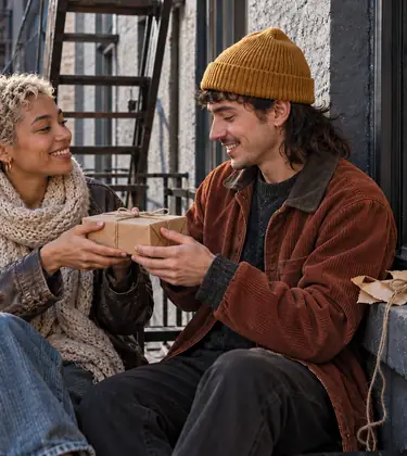 Two friends swap a kraft-wrapped gift on a city fire escape beside a rolling tray with a grinder, papers and a glass pipe