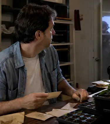 Man at a cluttered grow-room table with seed packets and seedlings looks toward shadowy figures at the door during a tense raid-like moment.