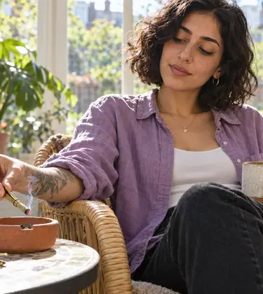 A woman in a sunroom holds the short roach of a joint in a brass roach clip over a terracotta ashtray
