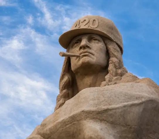 Stone monument wearing a carved stone 420 cap with a carved stone blunt in its mouth against a blue sky.
