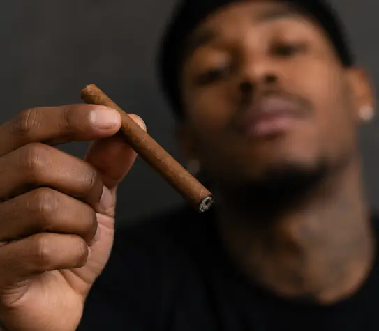 Professional athlete in dark sportswear holding a blunt in the foreground, with his face softly blurred in the background.