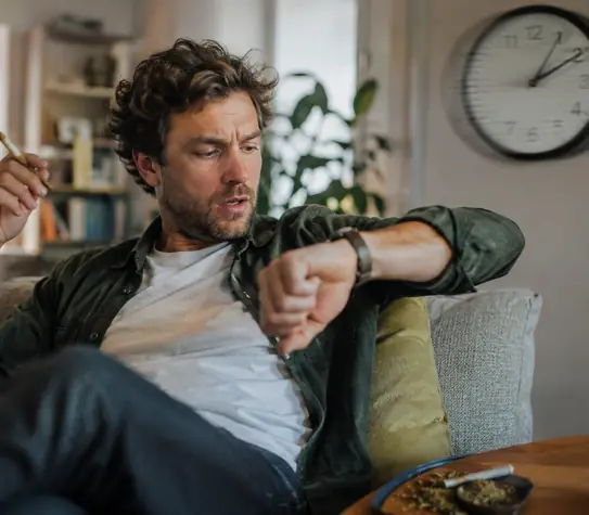 Man relaxing on a couch with a joint, checking his watch while a blurred wall clock suggests cannabis time distortion.
