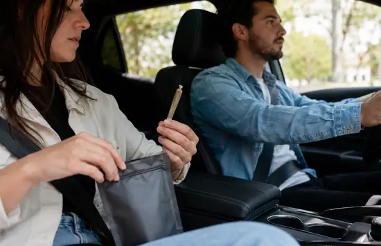Passenger holding an unlit cannabis pre-roll and resealable pouch inside a car while the driver focuses on the road.