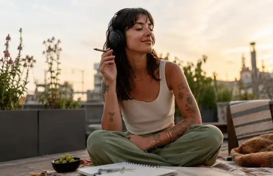 A relaxed woman wearing headphones sits on a rooftop blanket at sunset, holding a pre-roll with a sketchbook and snacks nearby.