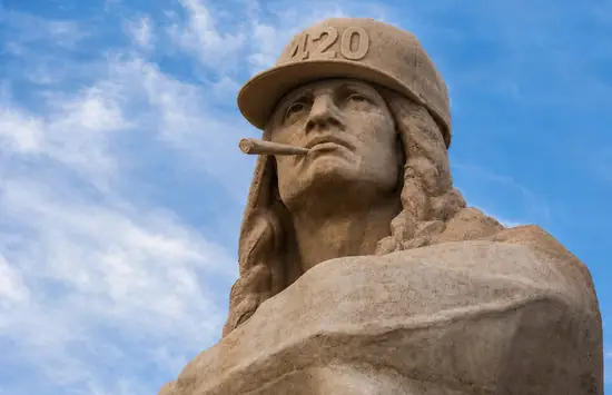 Stone monument wearing a carved stone 420 cap with a carved stone blunt in its mouth against a blue sky.
