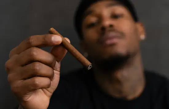 Professional athlete in dark sportswear holding a blunt in the foreground, with his face softly blurred in the background.