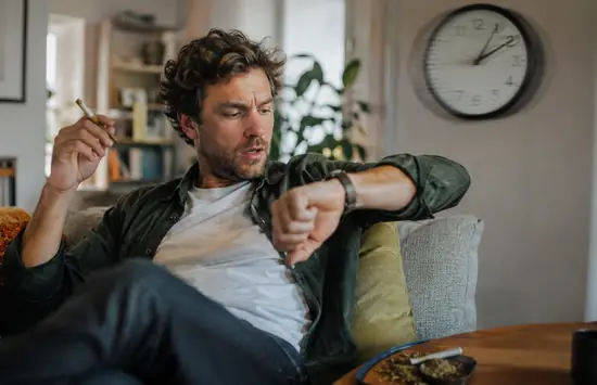 Man relaxing on a couch with a joint, checking his watch while a blurred wall clock suggests cannabis time distortion.