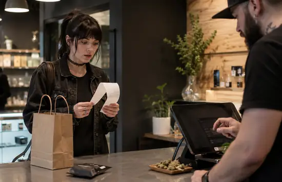 A surprised customer reads a long receipt at a modern cannabis dispensary checkout while a budtender operates the register.