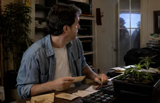 Man at a cluttered grow-room table with seed packets and seedlings looks toward shadowy figures at the door during a tense raid-like moment.