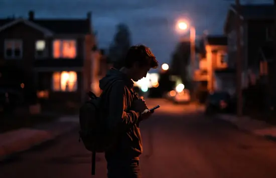 A young man in a hoodie with a backpack stands on a dimly lit residential street at dusk, checking his phone under streetlights